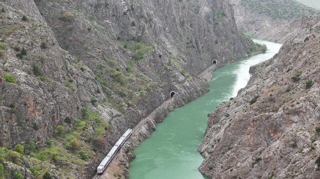 Aerial view of a passenger train passing through a deep rocky canyon next to a turquoise river.