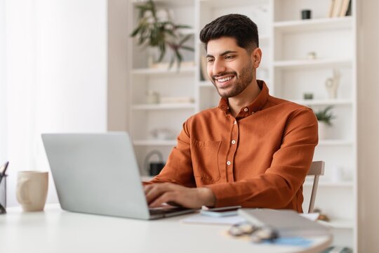 Smiling Young Caucasian Man Sitting At Desk Using Laptop In Modern Home Office. Focused On Work And Enjoying Productive Moment In Comfortable Environment, Promoting Business Concept.