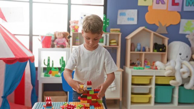 Boy stacking colorful plastic building blocks on table with both hands, toy tent and shelves visible in preschool classroom; creative concentration.