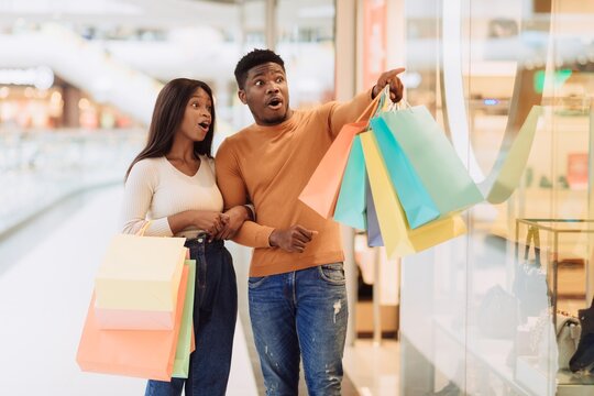 Joyful Black Couple, Young Adults, Shopping Together In Modern Mall. They Are Pointing At A Store Window, Filled With Colorful Shopping Bags And Exhibiting Excitement About Fashion And Style.