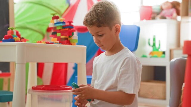 Boy holding a small plastic toy piece with his hand raised, carefully stacking colorful blocks on a low table in a building playroom; curiosity concentration.