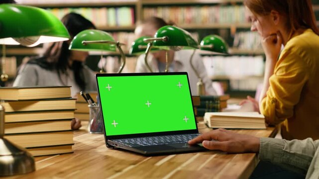 Chroma key green screen on laptop near student hand on wooden desk, using device with screen replacement at library, Isolated mockup on study table with soft focus on green lamps, with group study.