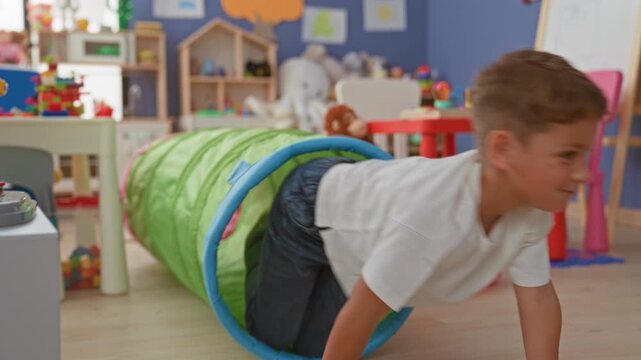 Boy crawls through green play tunnel between table and toys, reaching toward plush monkey in preschool classroom; curiosity play.