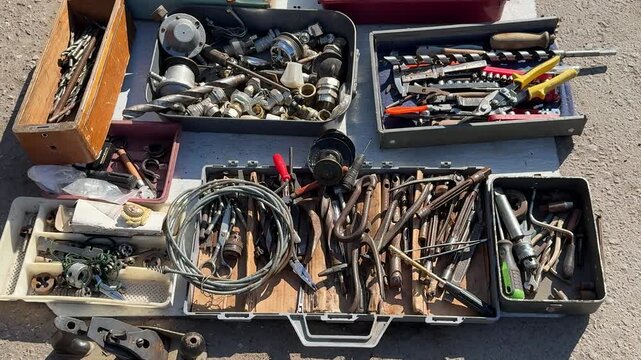 Used tools displayed on asphalt, assorted secondhand hand tools and small parts spread across open toolbox and plastic trays.