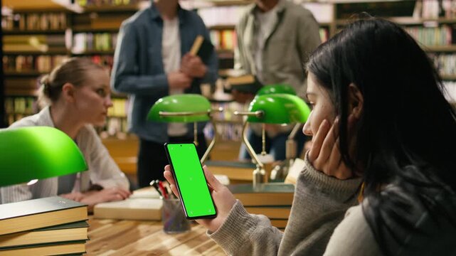 Vertical chroma key editable screen on smartphone held by person at library, green screen markers during study group, Mobile app mockup space with blurred green lamps, supporting screen replacement.