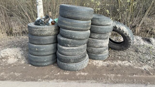 Stacked worn tires on gravel roadside. Birch trees background, columns of passenger car tires awaiting collection, weathered.
