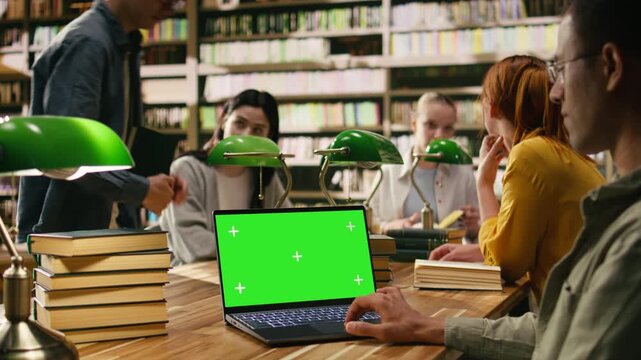 Chroma key isolated screen on laptop with student in over-the-shoulder shot, group study session in a library, Screen replacement with mockup near students at table, with coursework and green screen.