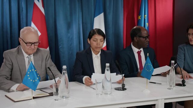 Multiethnic diplomats sitting at press conference table with European Union flags, discussing documents during summit. Clip is ideal for news broadcasts or global relations videos