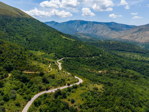 Aerial view of a winding mountain road snaking through lush green forests and rolling hills under a bright blue sky with scattered white clouds Albania.