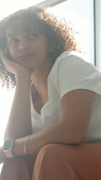 Vertical video: Camera revealing woman in white top, rust pants tilting head by window, smartwatch