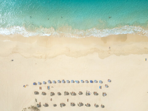 Aerial view of turquoise ocean waves washing onto golden sand with rows of beach umbrellas and people swimming at Santa Maria beach in Santa Maria, Sal, Cabo Verde.