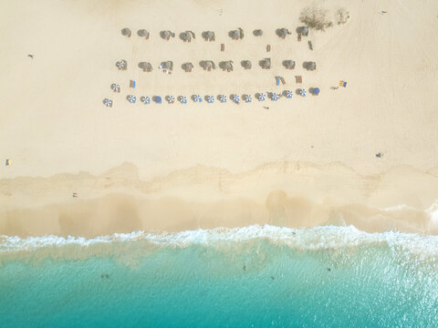 Aerial view of rows of beach umbrellas and sunbeds on the golden sand of Santa Maria beach with turquoise ocean waves and people swimming in Santa Maria, Sal, Cabo Verde.