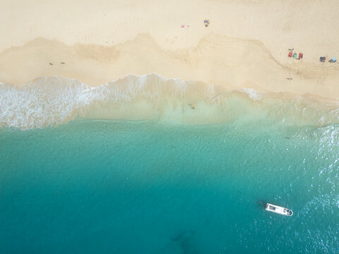 Aerial view of turquoise ocean waves washing onto the white sandy beach with a small boat in Santa Maria, Sal, Cabo Verde.