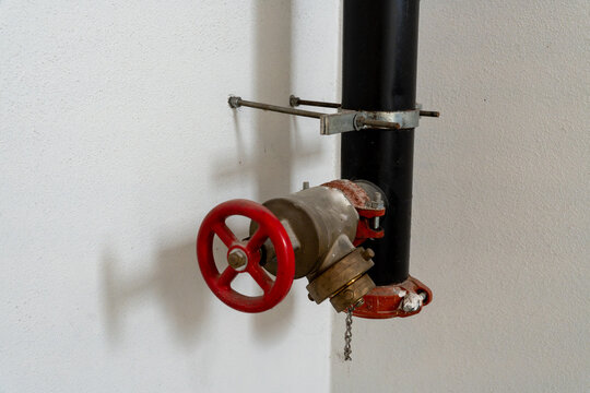 Horizontal 4K photo of a fire hose valve with a red handwheel mounted on a black pipe against a white textured wall