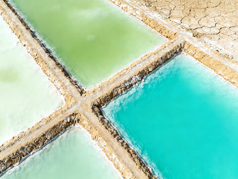 Aerial view of colorful salt evaporation ponds with varying shades of green and blue water, separated by earthen dikes in a dry landscape.