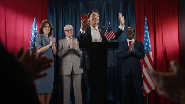 Young Hispanic female politician walking to podium, taking photo with smartphone while diverse colleagues applaud. Perfect for modern political campaign and election news videos wide shot