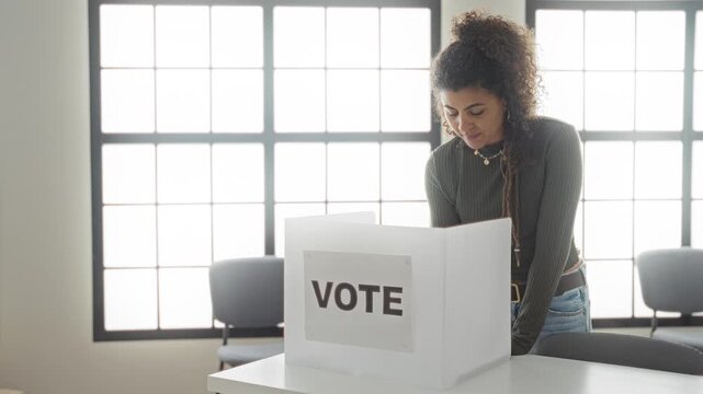 Woman marking a ballot at a voting booth inside a public building, leaning over the privacy screen to cast a vote; civic determination.