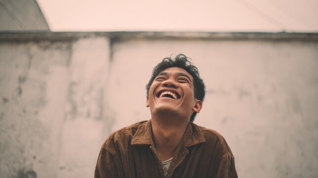 Young Asian man laughing joyfully outdoors against a wall