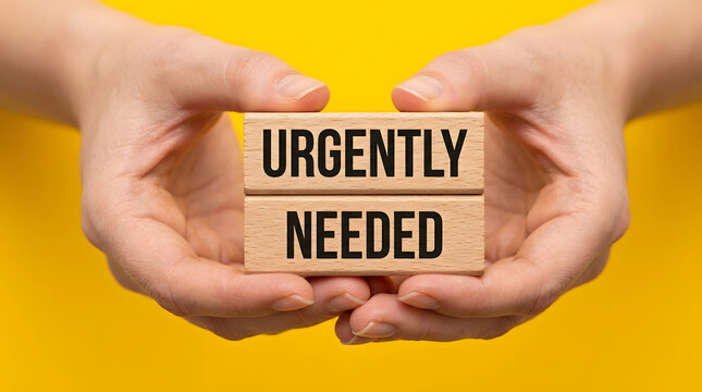 Hands holding wooden blocks with urgently needed text on yellow background