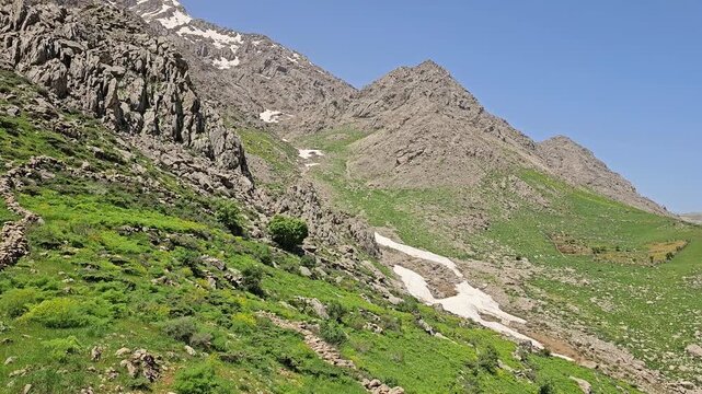 A green spring slope leads down into a rocky valley, where a snow patch lingers beneath distant snow-capped peaks
📍 Hawraman Takht, Kurdistan, Iran 