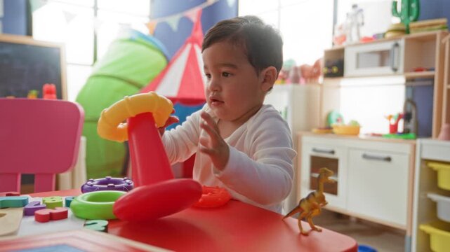 Boy stacking colorful plastic ring toy with hand on red table, reaching and placing rings while seated in building playroom; curiosity learning.