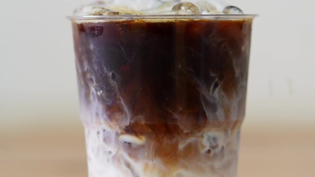 4K Close up of iced coffee latte with swirling dark espresso layers into fresh milk in clear plastic takeaway cup with ice cubes on wooden table, Refreshing morning cafe beverage concept.