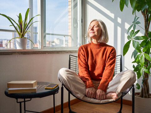 Content senior woman with closed eyes enjoying warm sunlight indoors, meditating or relaxing in peaceful home.