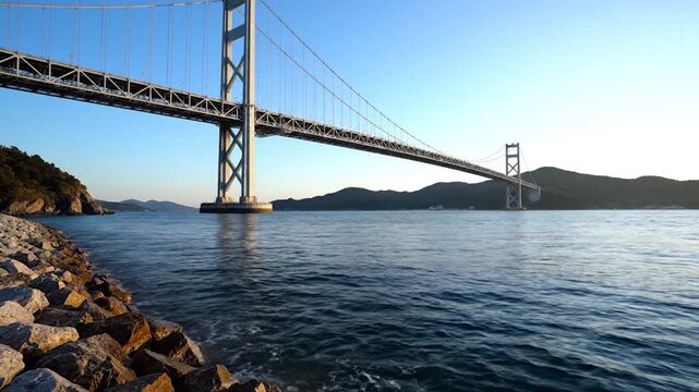 Large suspension bridge spanning the blue ocean under clear sky viewed from rocky shoreline