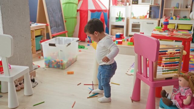 Little boy holding yellow cup and stepping over scattered crayons in a toy filled playroom inside a building; playful joy.