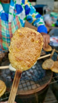 A street food vendor grills traditional sticky rice patties (Khao Jee) on bamboo skewers over a rustic charcoal stove at an outdoor market
