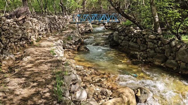 A rushing mountain stream with a small cascade, a rocky path alongside stone walls, and a blue bridge visible through lush spring trees ahead.
📍 Kamalla village, Hawraman Takht, Kurdistan 