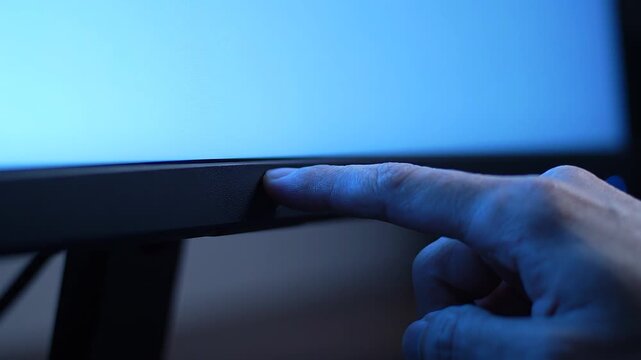 Finger presses power button on black monitor with blue screen glow