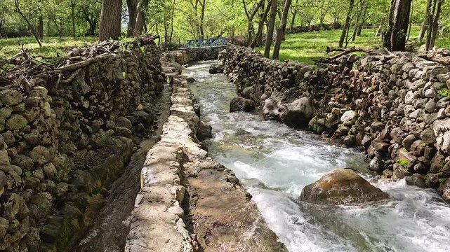 A spring valley with a rushing rocky stream and a narrow stone-walled water channel flowing side by side through lush green orchards, with a small bridge ahead.
📍 Kamalla village, Hawraman Takht, Kur
