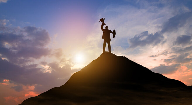 Silhouette of triumphant person standing on mountain peak holding trophy at sunset