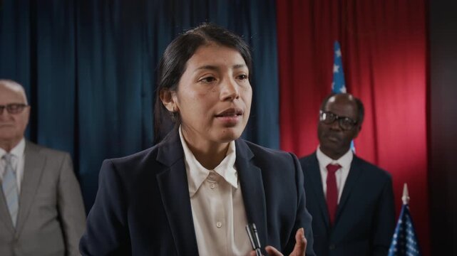 Young Hispanic woman politician speaking at podium, gesturing during press conference with diverse colleagues. Footage is suitable for political campaigns or news broadcasts