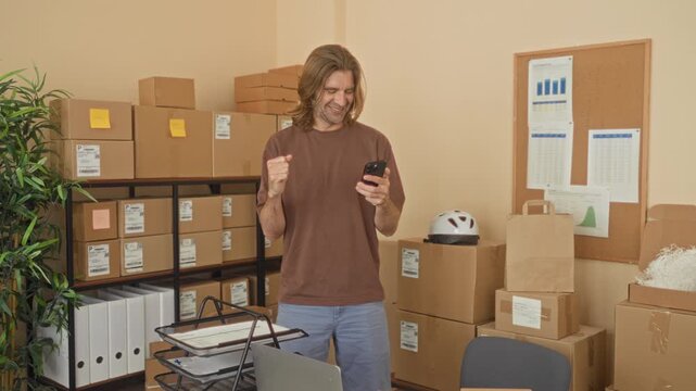 Man holding smartphone and doing a fist pump among stacked packages and shelves in a building; joy small business.