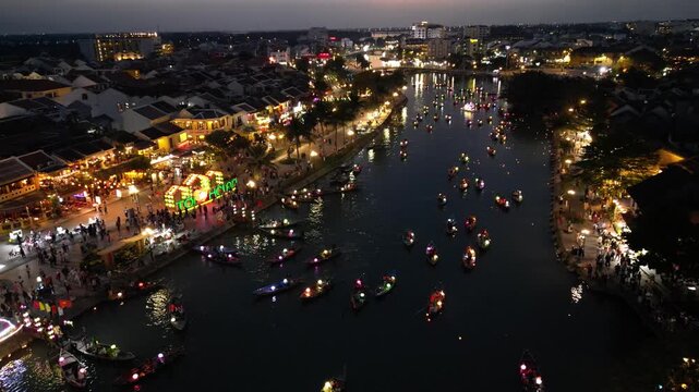 Aerial view of traditional lantern boats floating on the Hoai River at night with illuminated historic buildings in Hoi An, Da Nang, Vietnam.