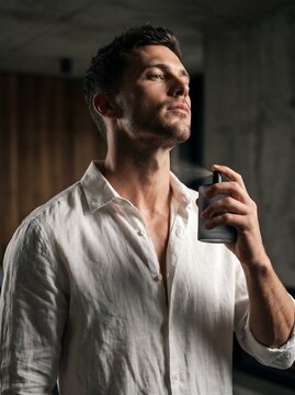 Fit young man in an open white linen shirt spraying a fresh masculine cologne bottle on his neck, conveying seductive sensory appeal and daily care