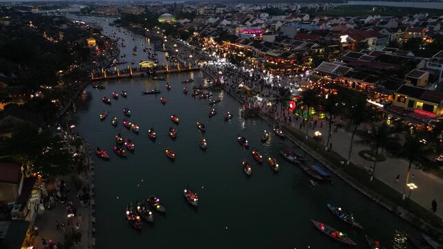 Aerial view of numerous lantern boats floating on the Thu Bon River at night with crowds of people along the illuminated banks of the ancient town in Hoi An, Da Nang, Vietnam.