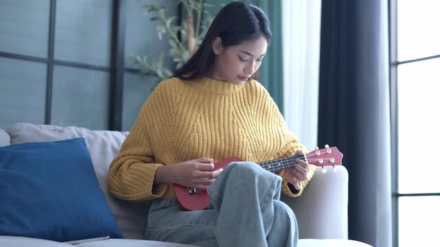 Asian woman in sweater playing ukulele in living room