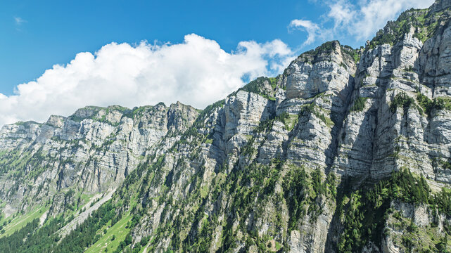 A scenic view of the majestic Swiss Alps mountain range in Switzerland, featuring rugged limestone cliffs, lush green forests, and a bright blue sky with soft white clouds.