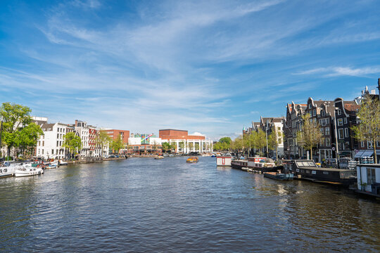 A scenic view of the Amstel River featuring historic canal houses and houseboats in Amsterdam, Netherlands. The bright sunny day highlights the classic Dutch urban architecture.
