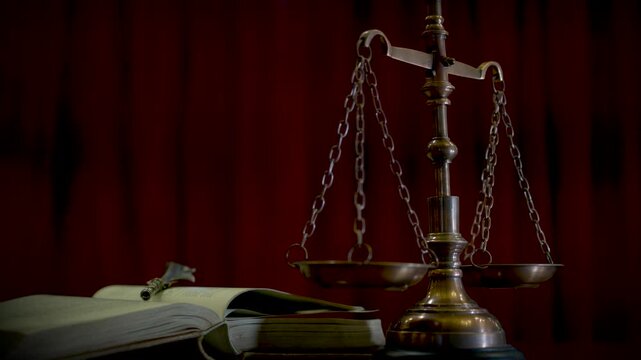 A brass scale of justice displayed against a deep red curtain background on a wooden surface evoking a classic courtroom atmosphere.