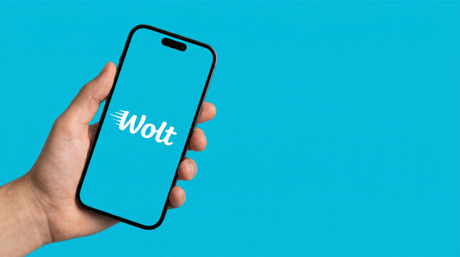 Male hand holding a smartphone iPhone with Wolt food delivery app logo on the screen. Modern mobile application interface mockup on light blue background with copy space for text and brand logo.