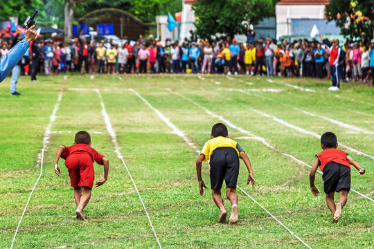 Schoolboys starting barefoot running race with referee firing pistol. Back view of young schoolboys sprinting barefoot from the starting line on a grass field track at outdoor sports day event.