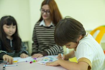 Fototapeta premium A smiling female teacher watches over a young Asian girl drawing with markers and a young boy exploring with a toy microscope during a fun learning activity in a bright kindergarten.