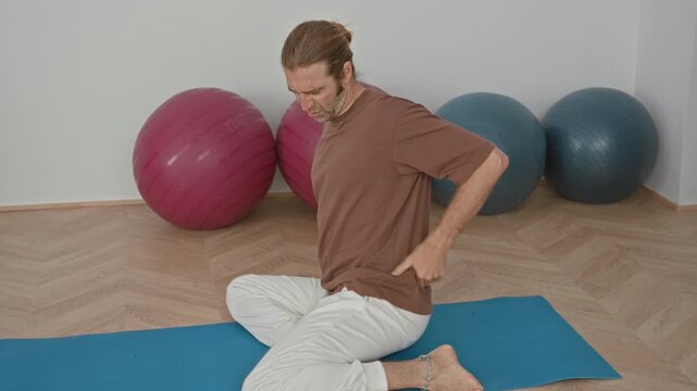 Young blond man on blue yoga mat holds lower back and winces beside pink and gray exercise balls in studio; discomfort recovery.