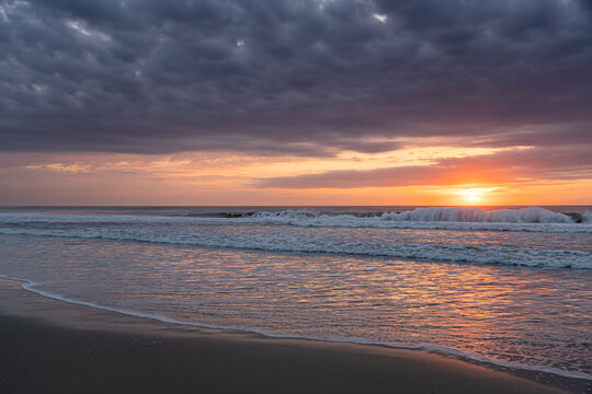 Dramatic sunrise over breaking waves on the beach in Corolla, North Carolina. Dark storm clouds contrasting with a vibrant orange sun on the Outer Banks shoreline