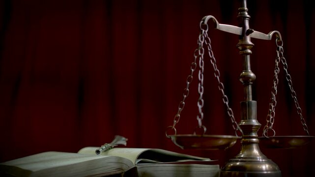 A brass scale of justice figurine placed on a wooden surface with a rich dark red curtain backdrop and soft warm atmospheric lighting.