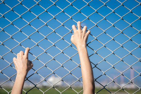Closeup of a human hand gripping a chain link fence as if trying to escape, representing border control, imprisonment, human trafficking, and the fight against restricted movement and injustice.